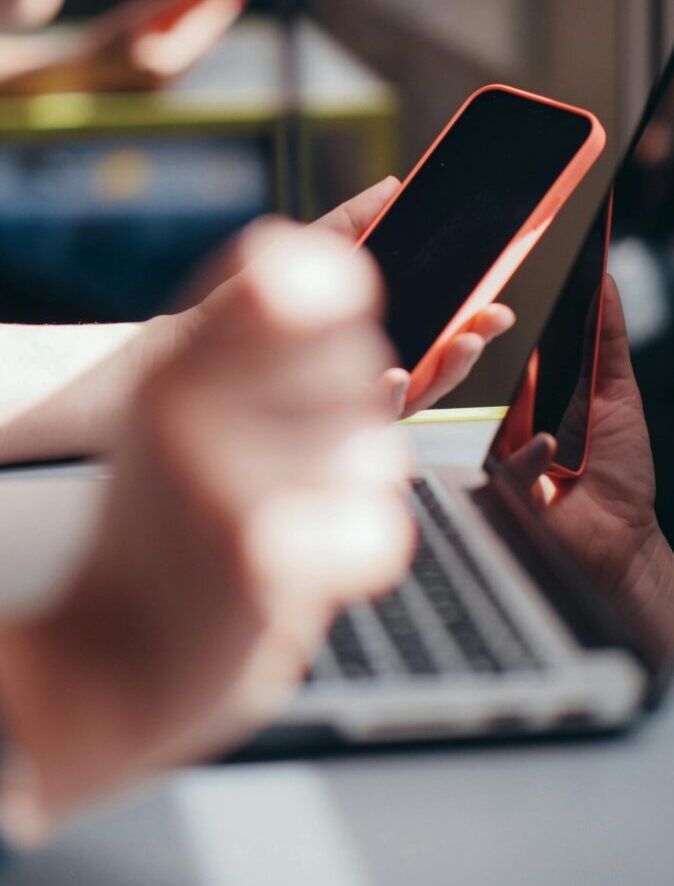 Hands holding smartphone near laptop, showcasing multitasking and technology use.